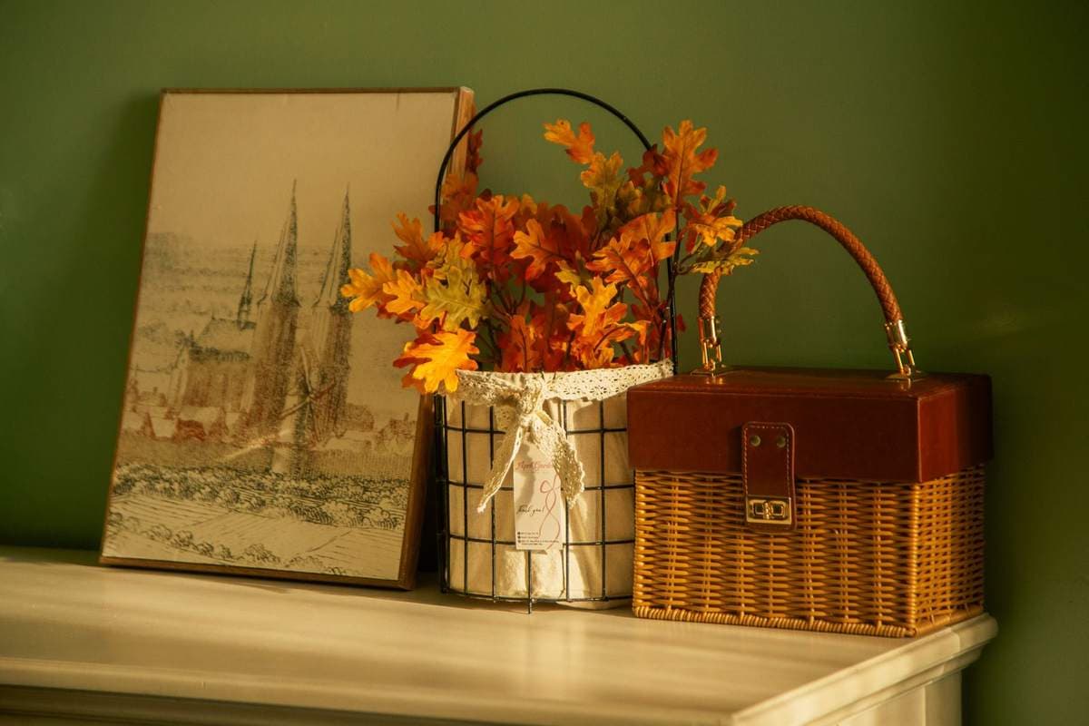 Framed artwork leaning on a mantel beside a basket of orange autumn leaves and a wicker case