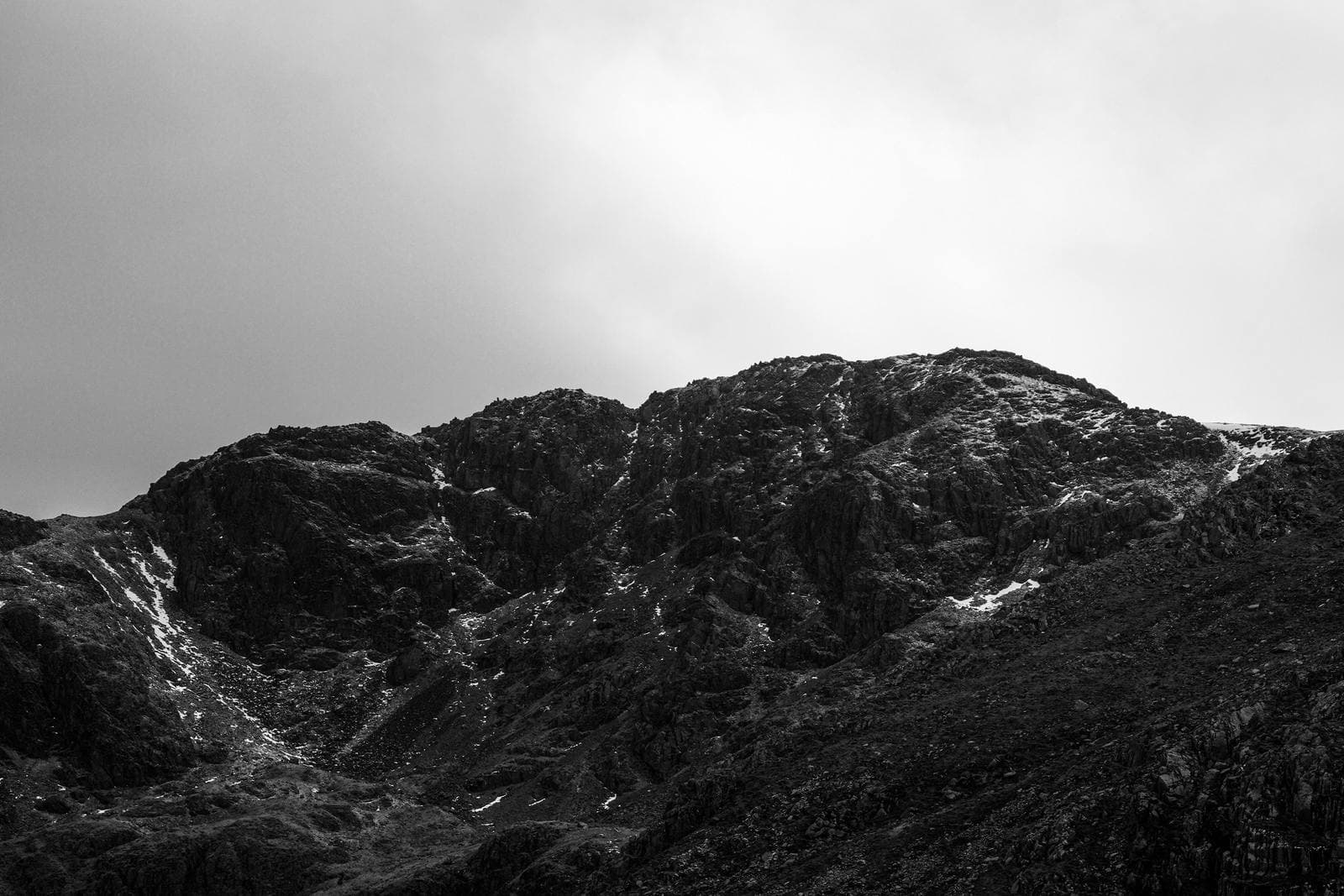 A black-and-white photograph of a rugged mountain ridge beneath a cloudy sky, showing the tonal range of monochrome landscape photography.