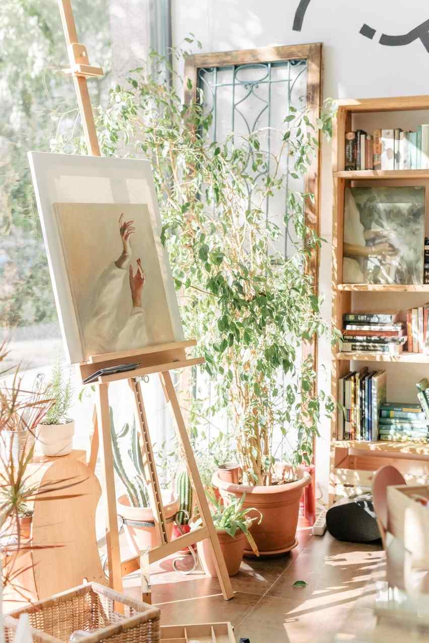 A painted canvas on a wooden easel in a sunlit artist studio with plants and bookshelves