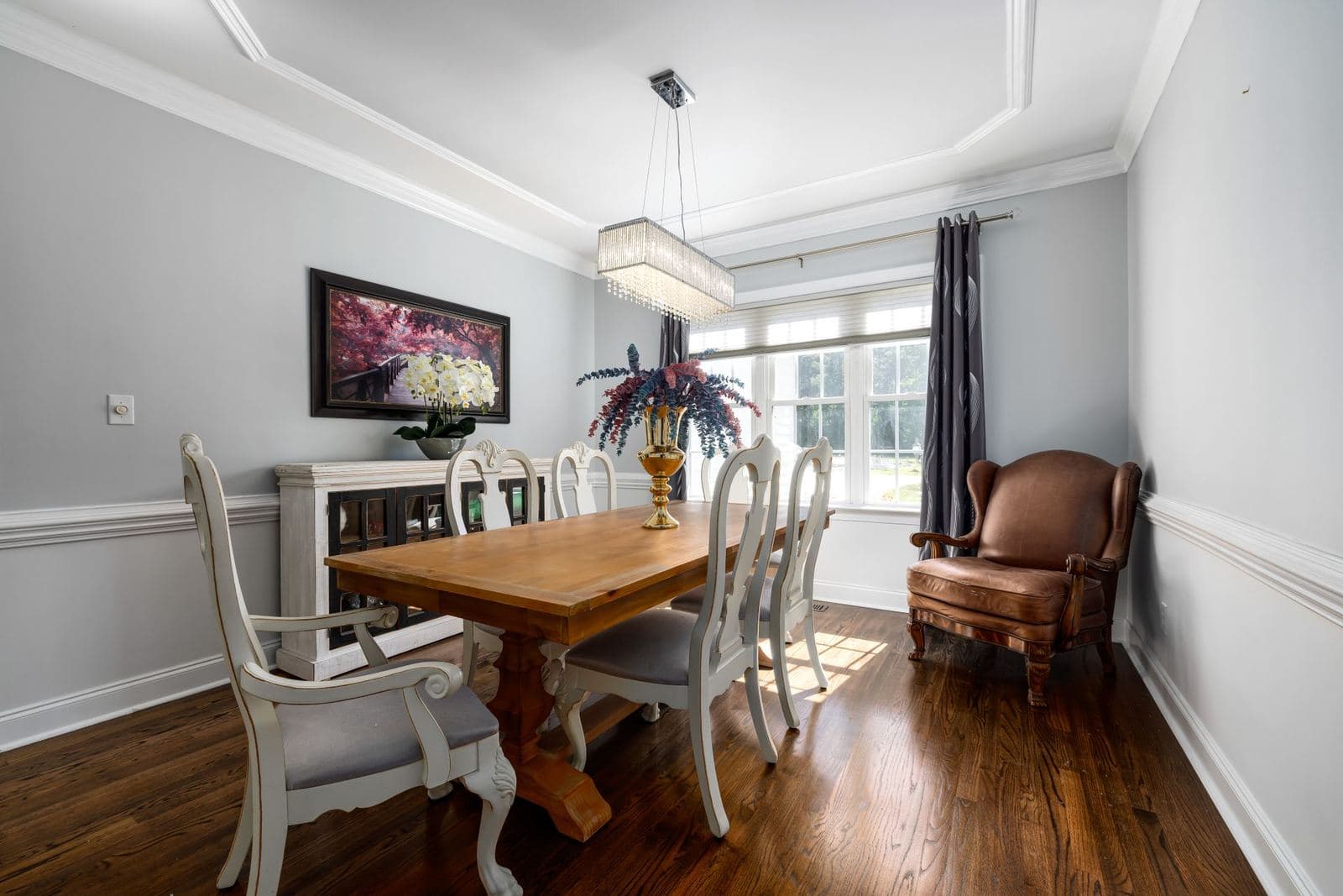 Bright dining room with a wooden table and white chairs, a framed floral picture above the sideboard and a leather armchair in the corner.