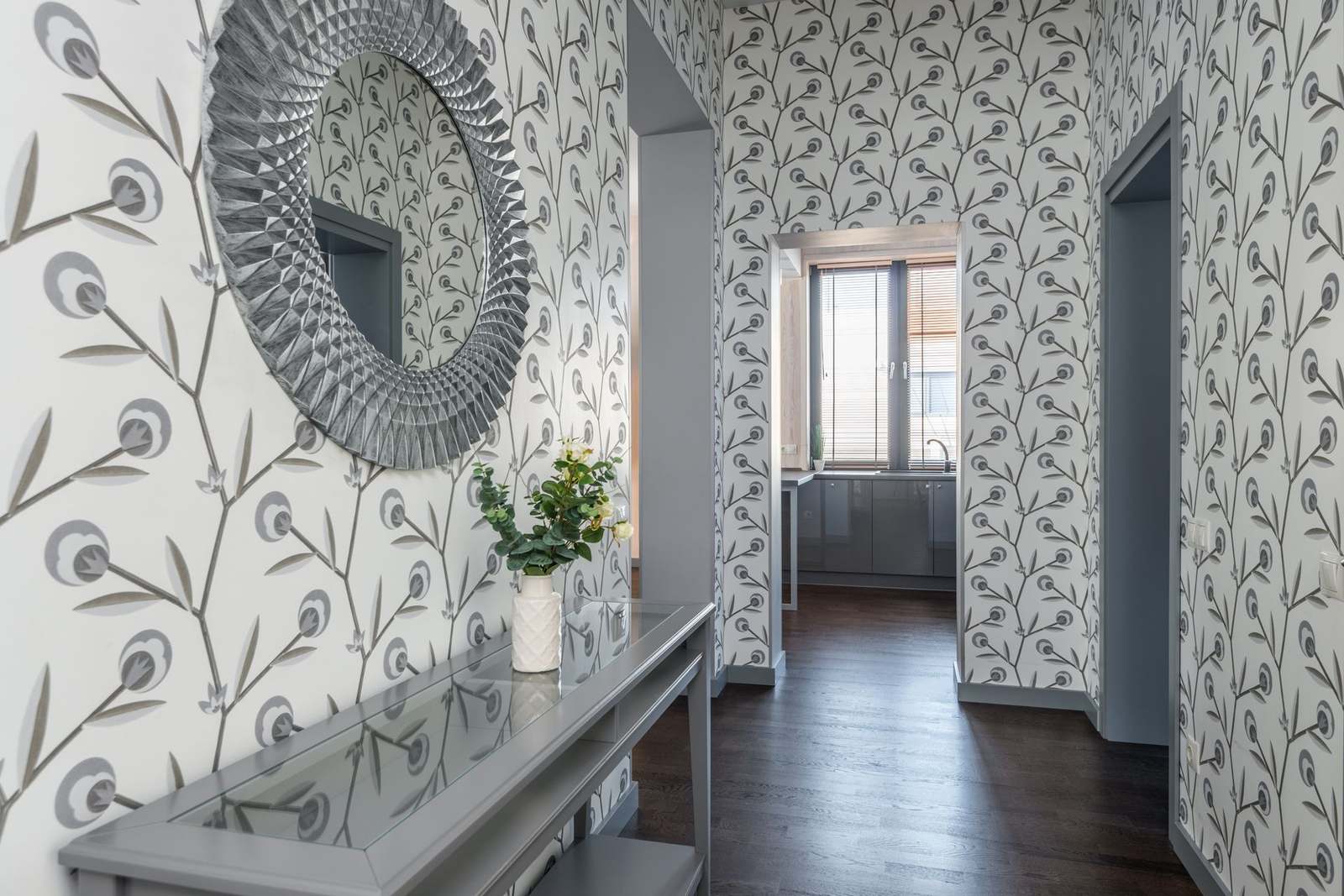 Hallway with botanical wallpaper, a grey console table with a vase of greenery, and a large round textured mirror.