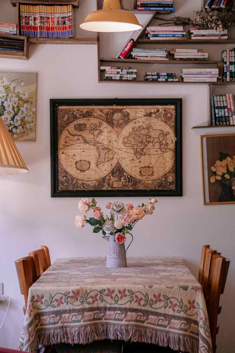 Framed antique world map hanging above a dining table with a vase of roses, surrounded by bookshelves