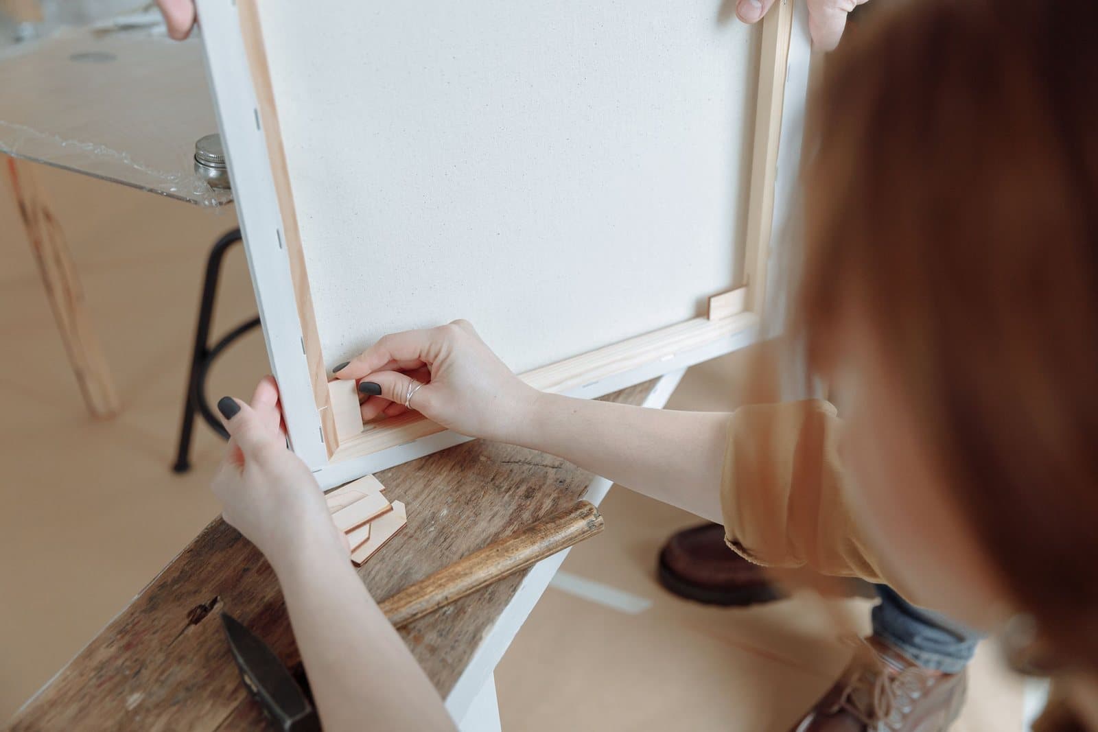 Hands fitting a wooden corner key into the back of a canvas on its stretcher frame at a workbench.