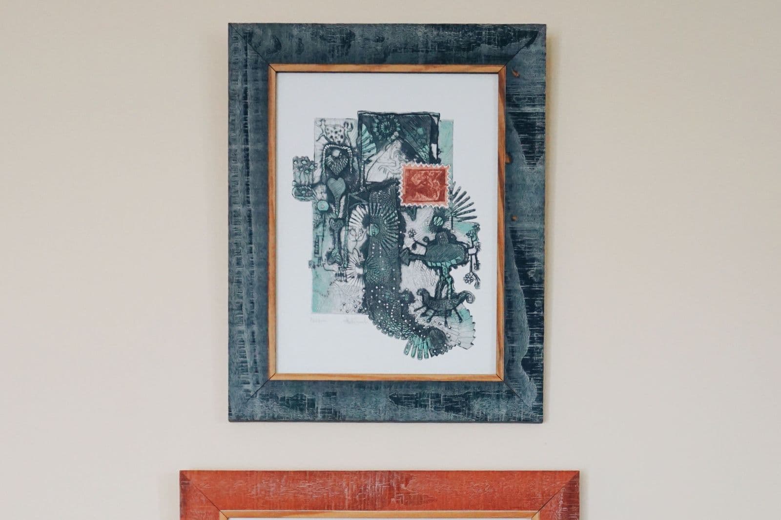 Two framed artworks in contrasting frame finishes — a dark teal marbled frame above a rust-red frame — hanging on a cream wall beside a houseplant.