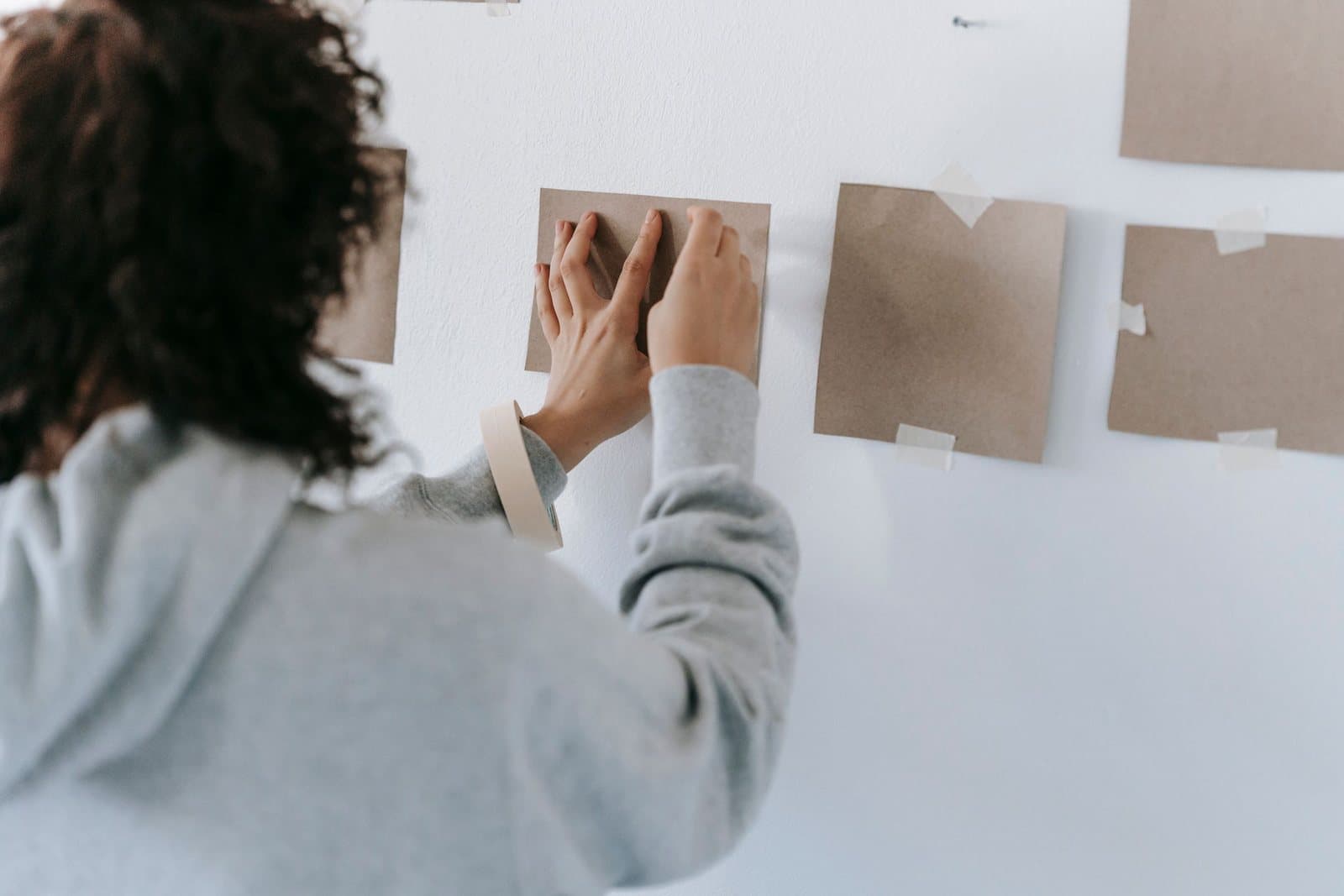 A woman taping brown paper templates to a white wall to measure out the size and placement of artwork before hanging it.