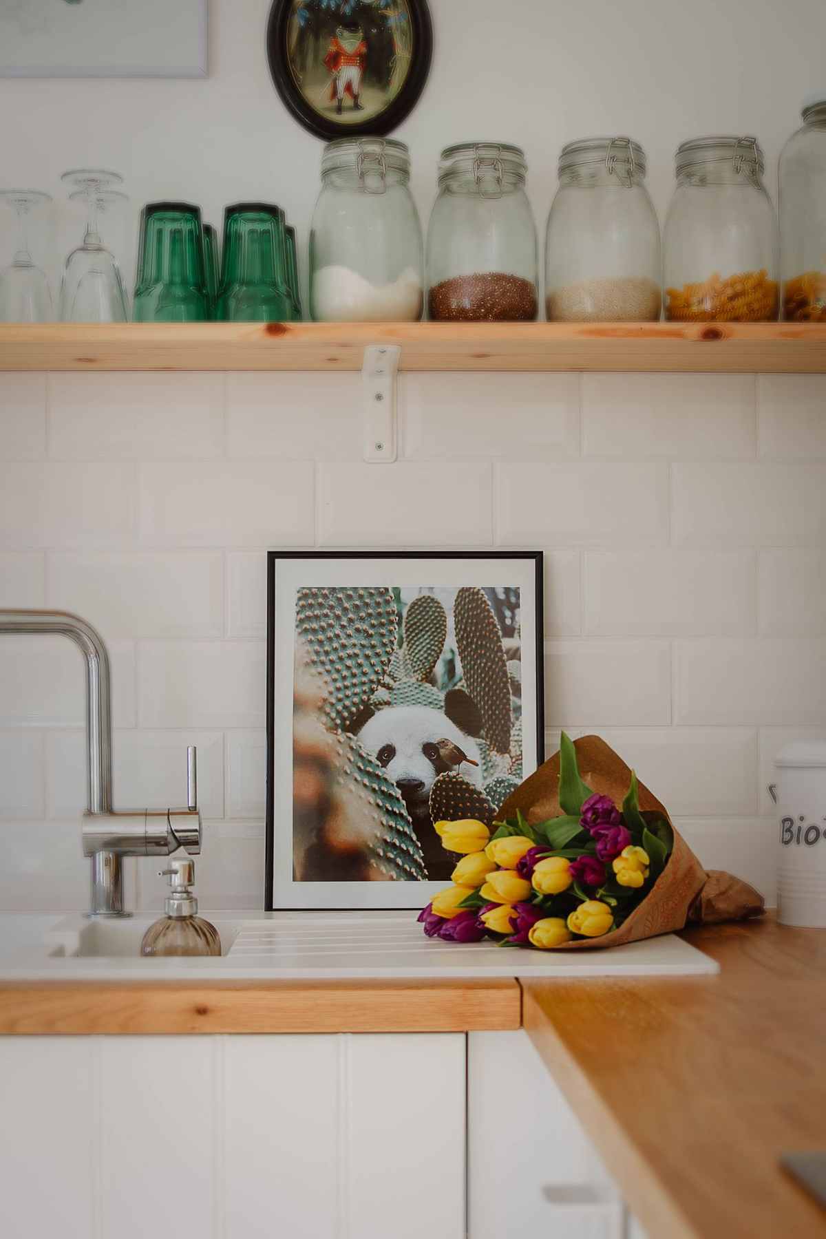 A framed picture leaning on a kitchen counter by the sink, with glass storage jars on a wooden shelf above and a bouquet of tulips beside it