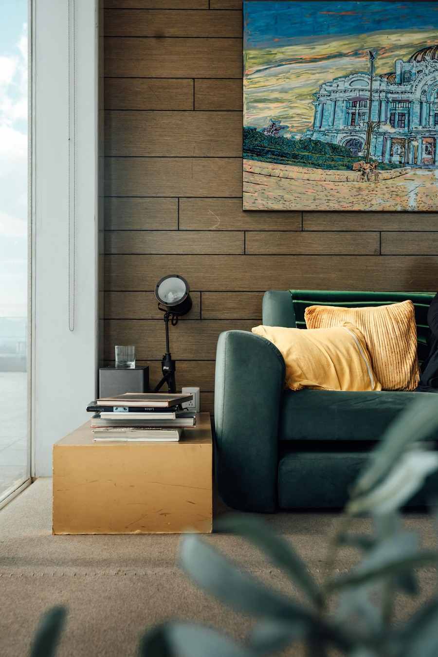 A large colorful painting of a grand building hanging on a wood-panelled wall above a green sofa with yellow cushions