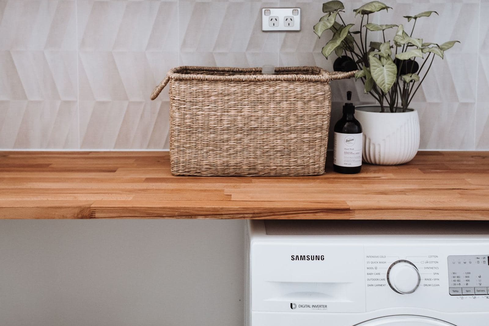 Bright laundry corner with a washing machine under a wooden counter holding a wicker basket and a potted plant.