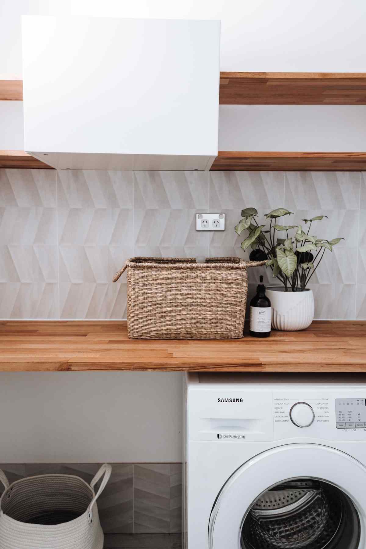 A laundry room with a washing machine under a wooden counter holding a wicker basket and a potted plant, with open wall space above