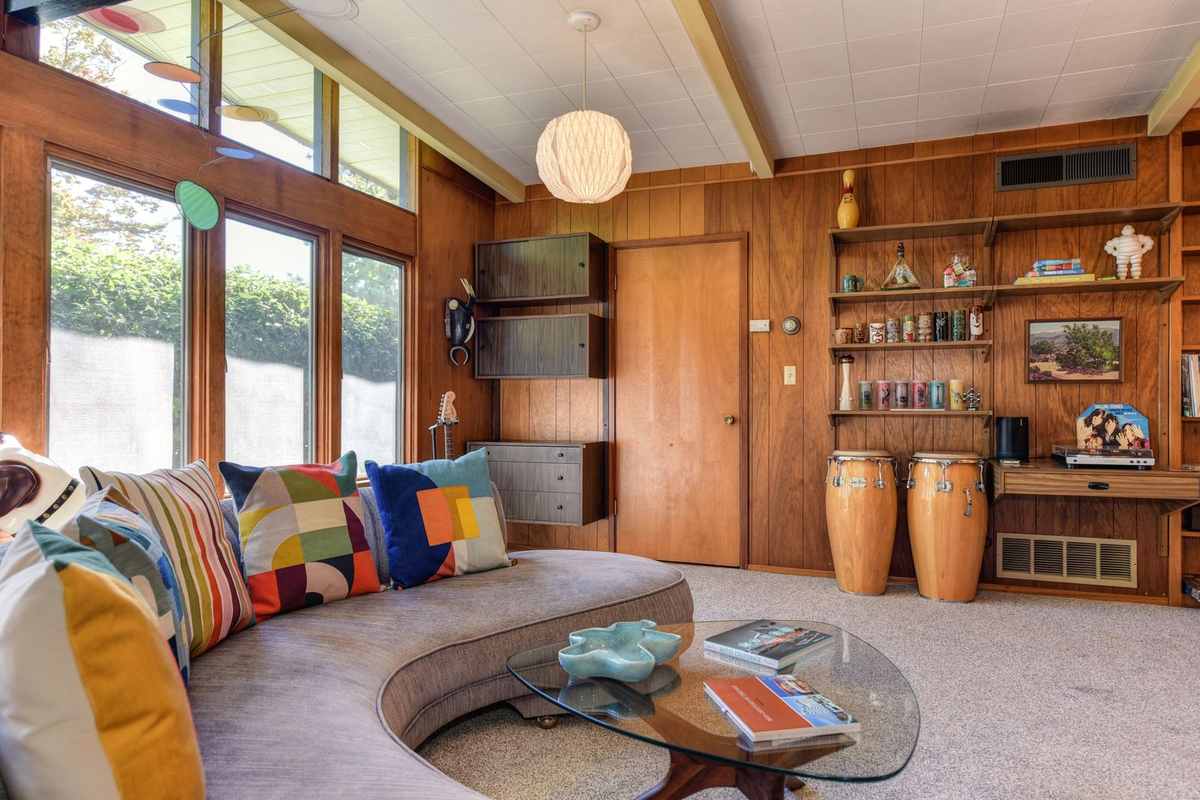 A mid-century modern living room with wood-panelled walls, a curved sofa, colorful geometric cushions and a small framed landscape on a shelf