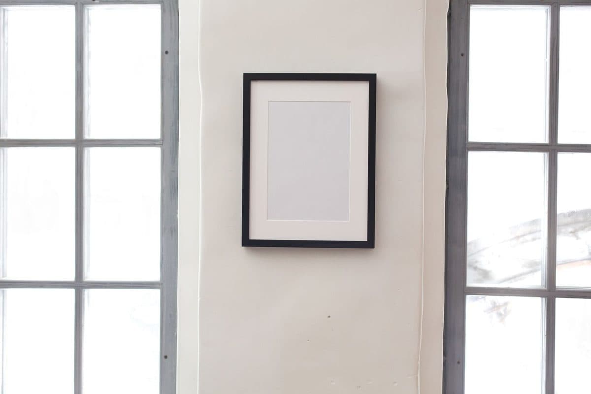 A single black frame with a wide white mat hanging on a white wall between two tall windows