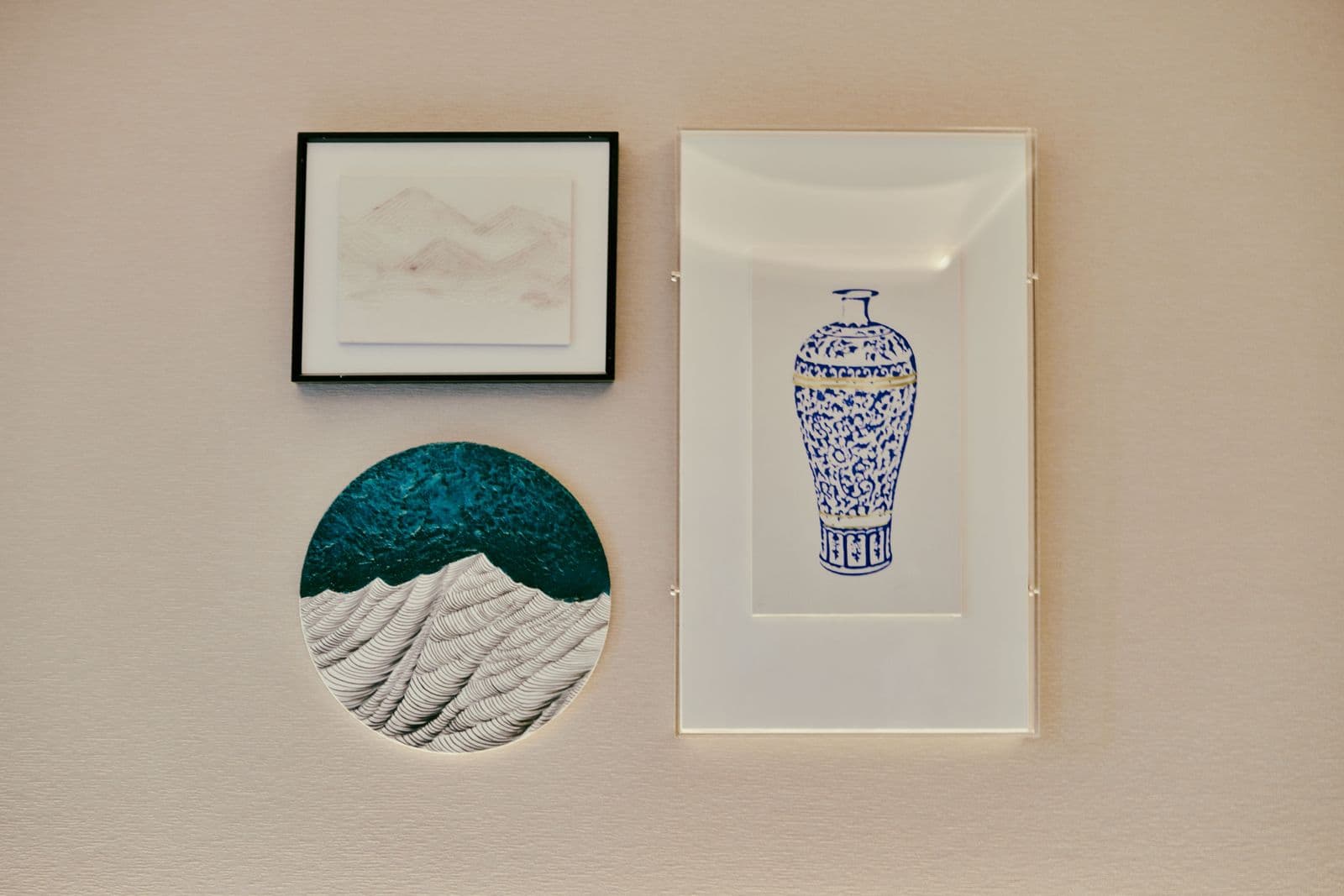A textured beige wall displaying three different wall art formats side by side: a small framed print, a round canvas with a wave design, and a tall framed print of a blue-and-white vase.