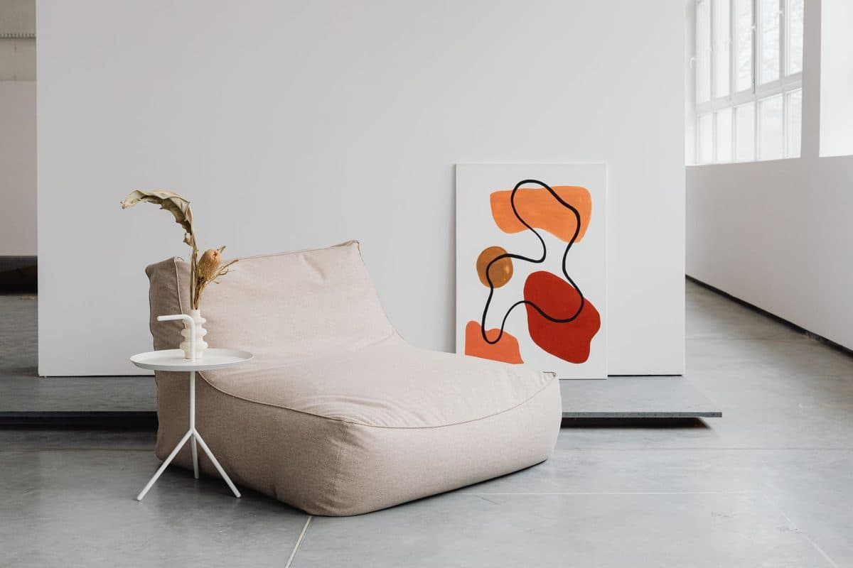 Bold orange and red abstract canvas leaning against a white wall in a neutral room with a beige lounge chair