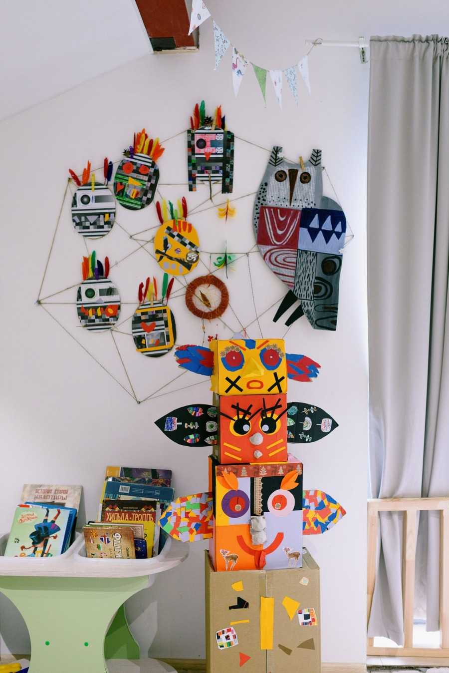 A childrens room wall covered with colorful handmade owl and character decorations above stacked cardboard figures and books