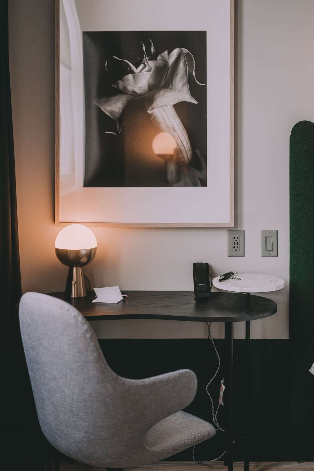 A large framed black-and-white floral photograph with deep shadows hanging above a desk and a lit table lamp