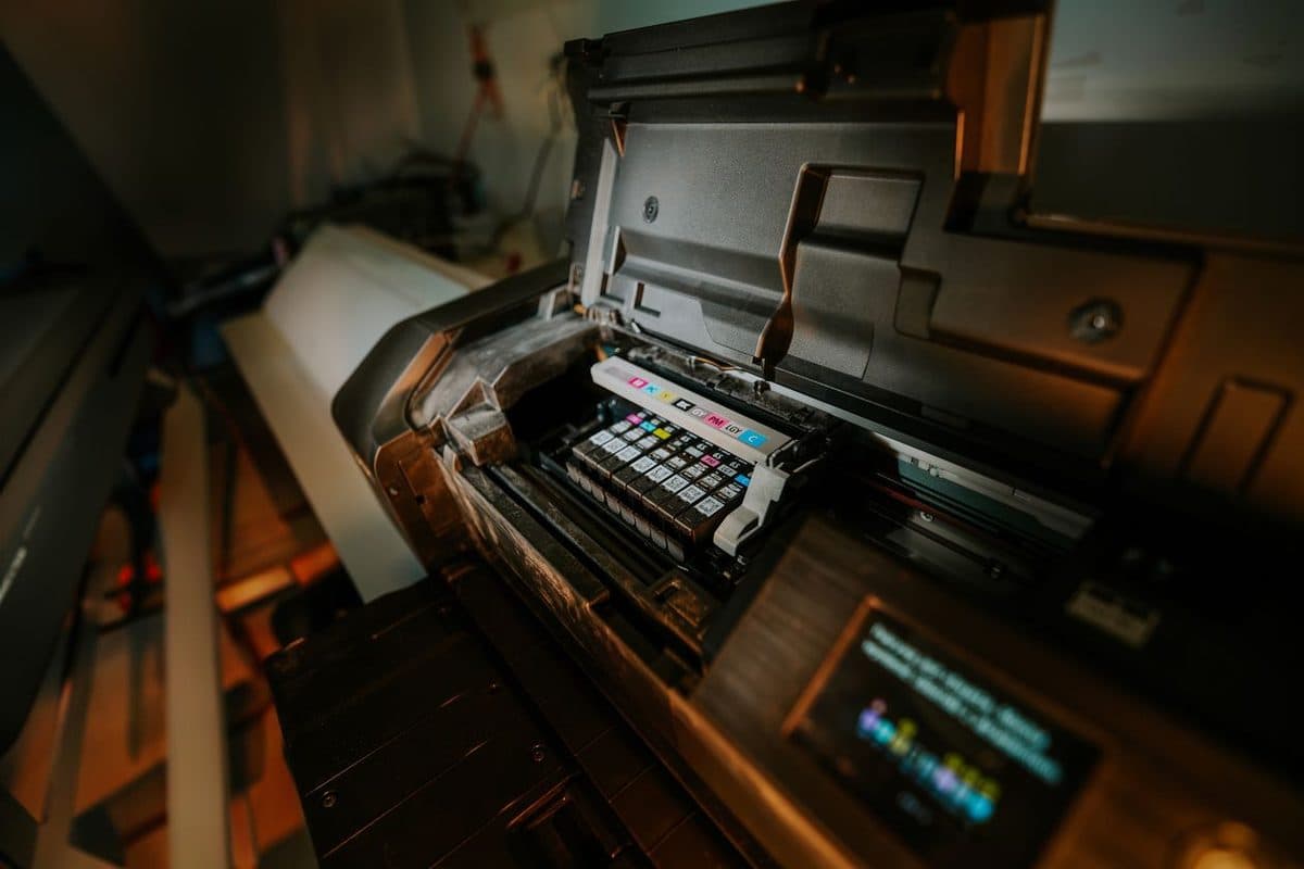 A large-format inkjet printer with its cover open, showing rows of colour ink cartridges and a roll of paper