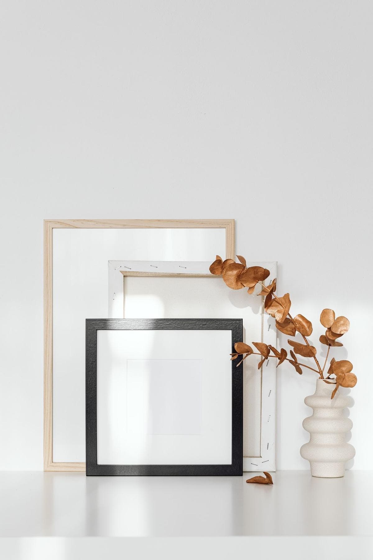 Empty frames leaning against a white wall, the front black frame holding a sheet with a wide white mat, beside a vase of dried eucalyptus