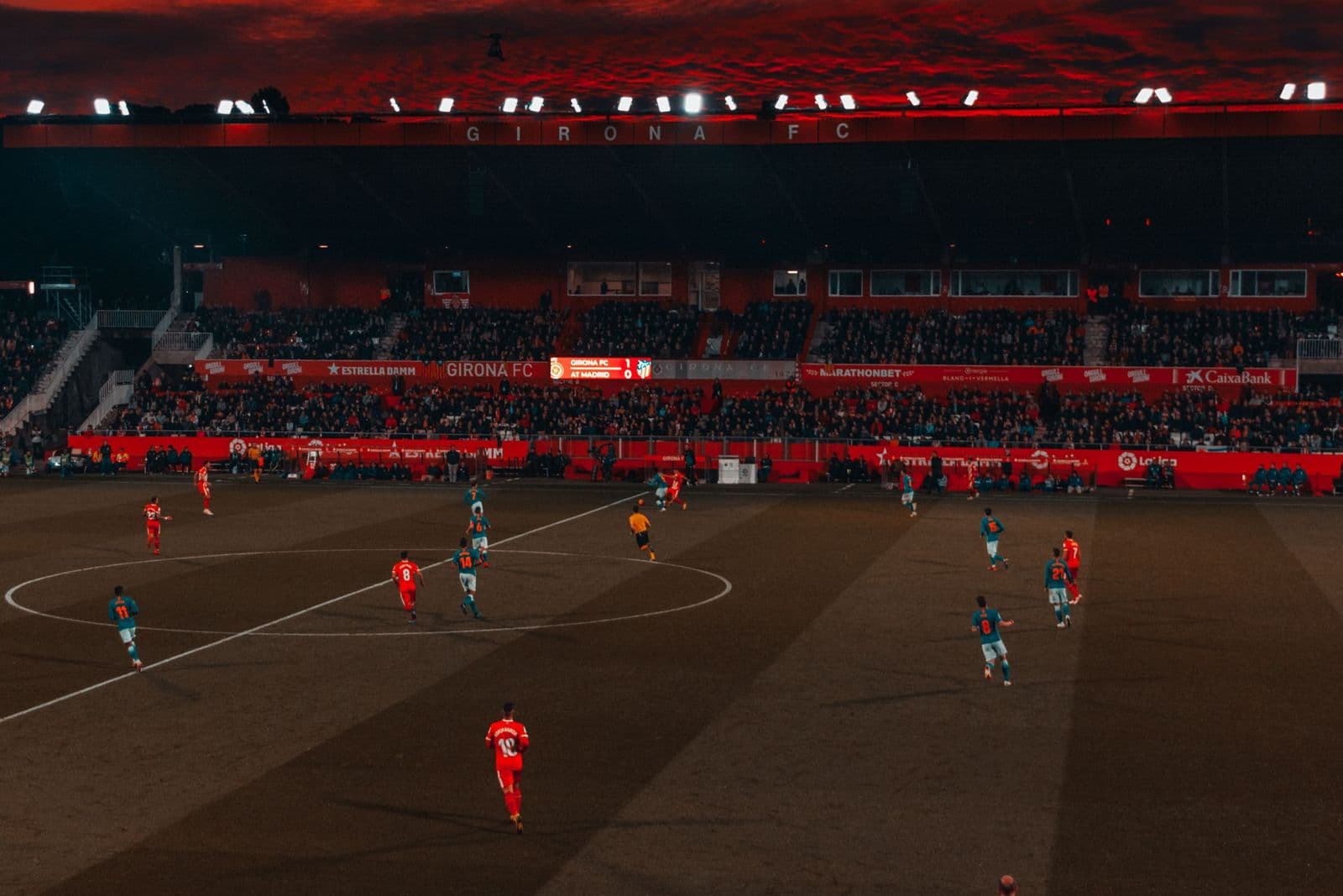 A soccer match under floodlights in a packed stadium beneath a dramatic red sunset sky.