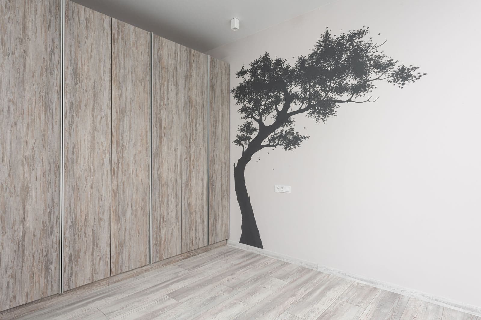 A black tree-shaped wall decal applied directly to the white wall of an empty room with light wood flooring and built-in wardrobes.