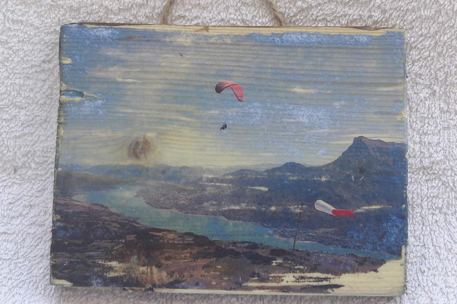 A landscape photograph of a lake, mountains, a paraglider and a windsock transferred onto a wooden panel, the wood grain and a knot showing through the image, the panel hanging by a twine loop